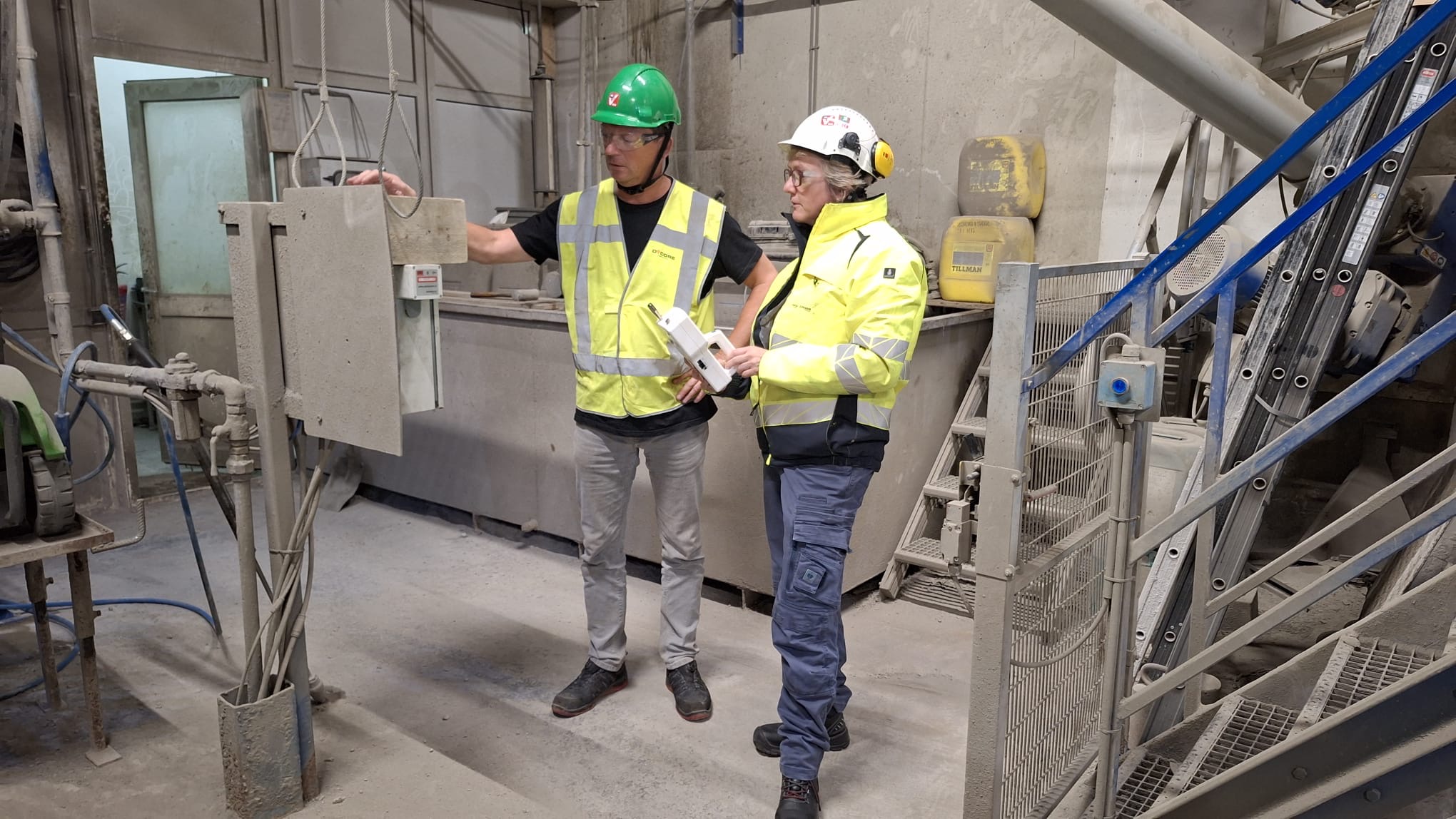 Arbeidshygiënist Imke Breedveld (rechts) met een handheld stofmonitor in overleg met Hoofd Technische Dienst Jos de Laat (links) bij een stationaire stofsensor op de betonmengerij van Dycore Breda. Foto: VGM Breed